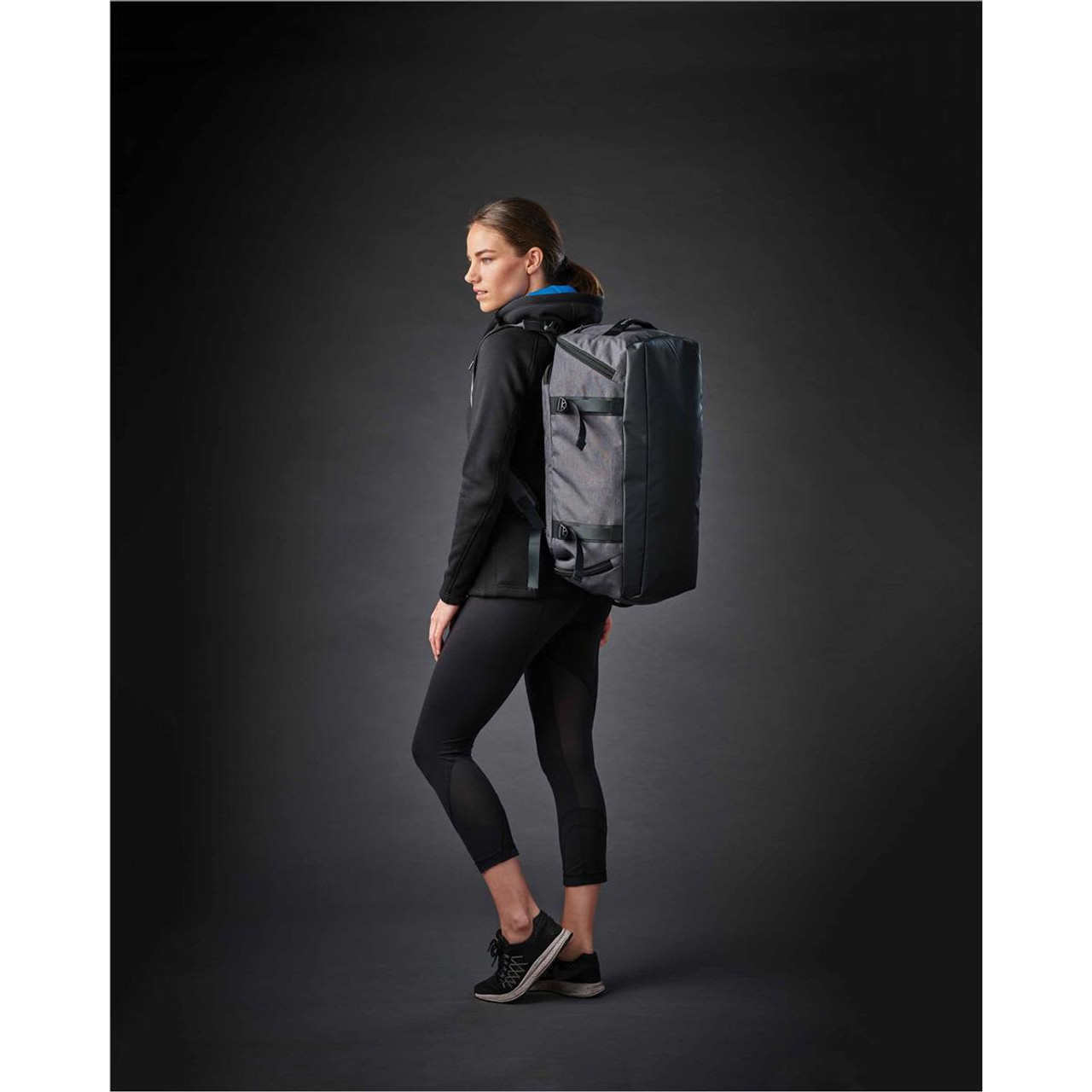 A woman in athletic wear stands sideways, carrying a grey Equinox 30 duffel bag with a logo.