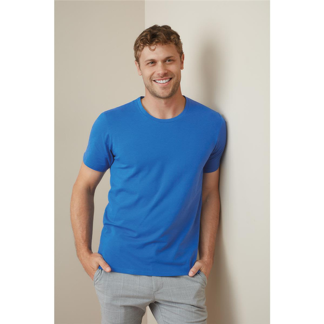 A blue crew neck t-shirt for men, featuring short sleeves and a casual design, with a model standing against a wall. A blue crew neck t-shirt for men, featuring short sleeves and a casual design, with a model standing against a wall.