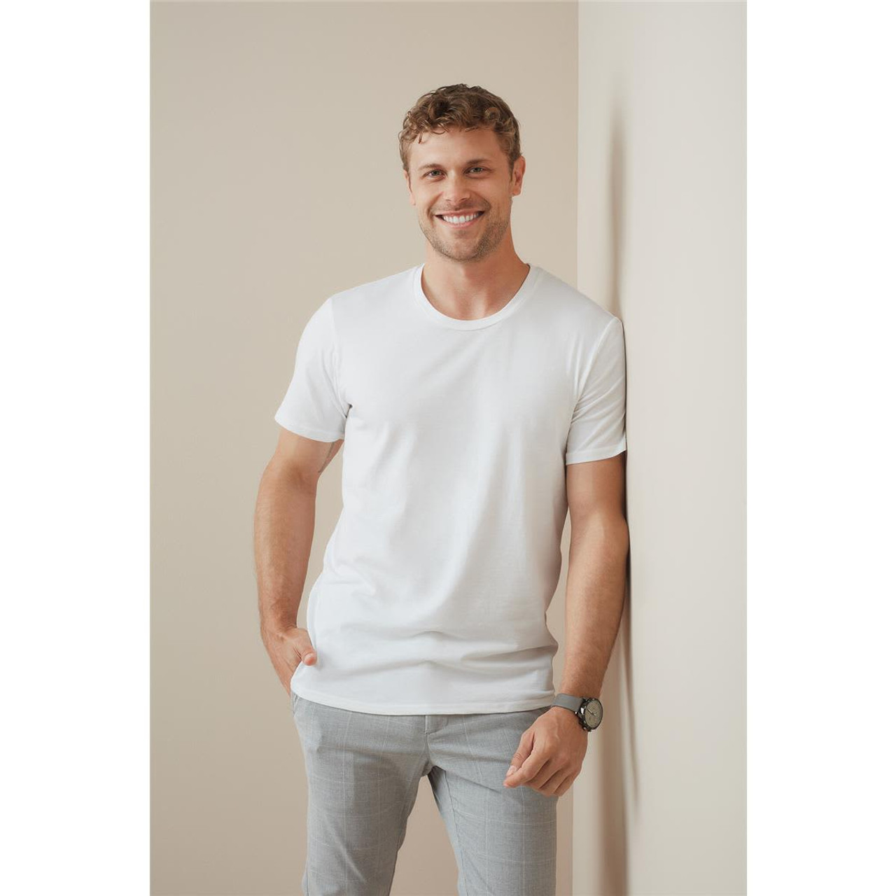 A man in a white crew neck t-shirt leans against a wall, smiling and wearing light grey pants. A man in a white crew neck t-shirt leans against a wall, smiling and wearing light grey pants.