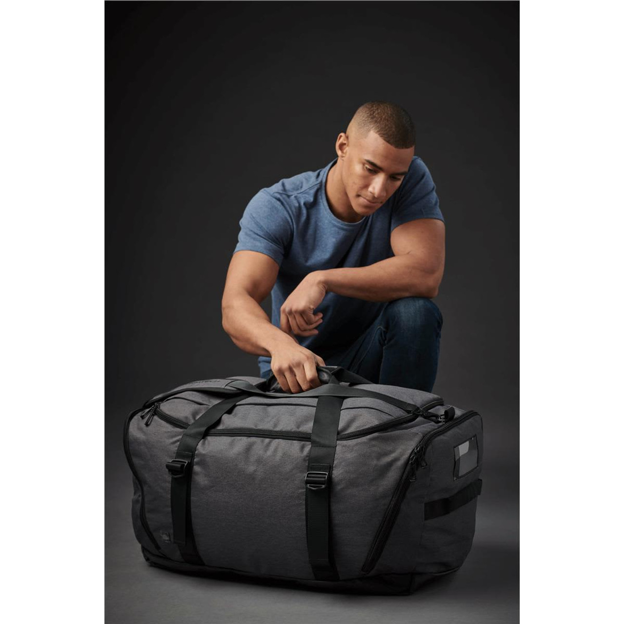 A grey duffle bag with green straps sits on the floor next to a man. The bag has a logo and a zippered compartment.