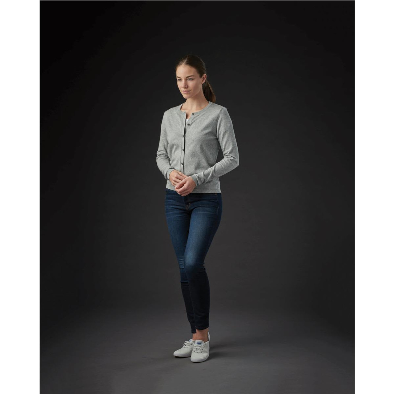 A grey women's cardigan with buttons, worn over fitted jeans and paired with white sneakers, displayed against a dark background.
