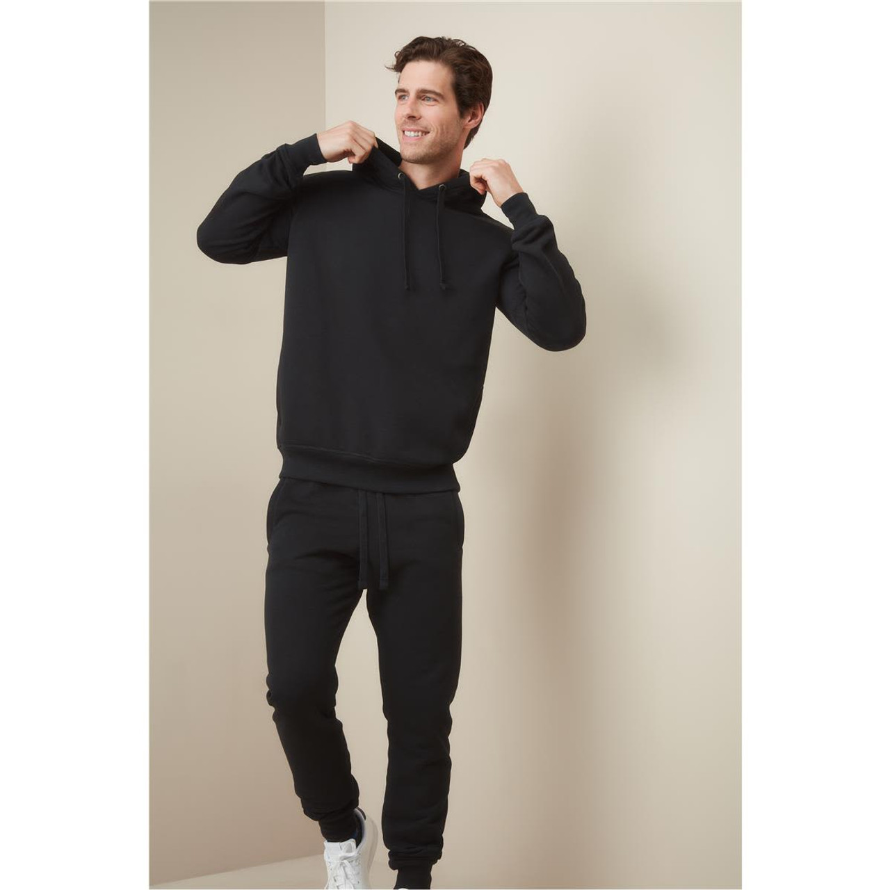 A man in black recycled unisex sweatpants and a matching hoodie, adjusting his hood, against a neutral background.