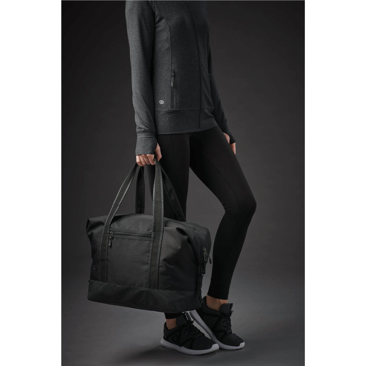 A black duffle bag with sturdy handles, held by a person wearing a dark outfit and sneakers. A black duffle bag with sturdy handles, held by a person wearing a dark outfit and sneakers.