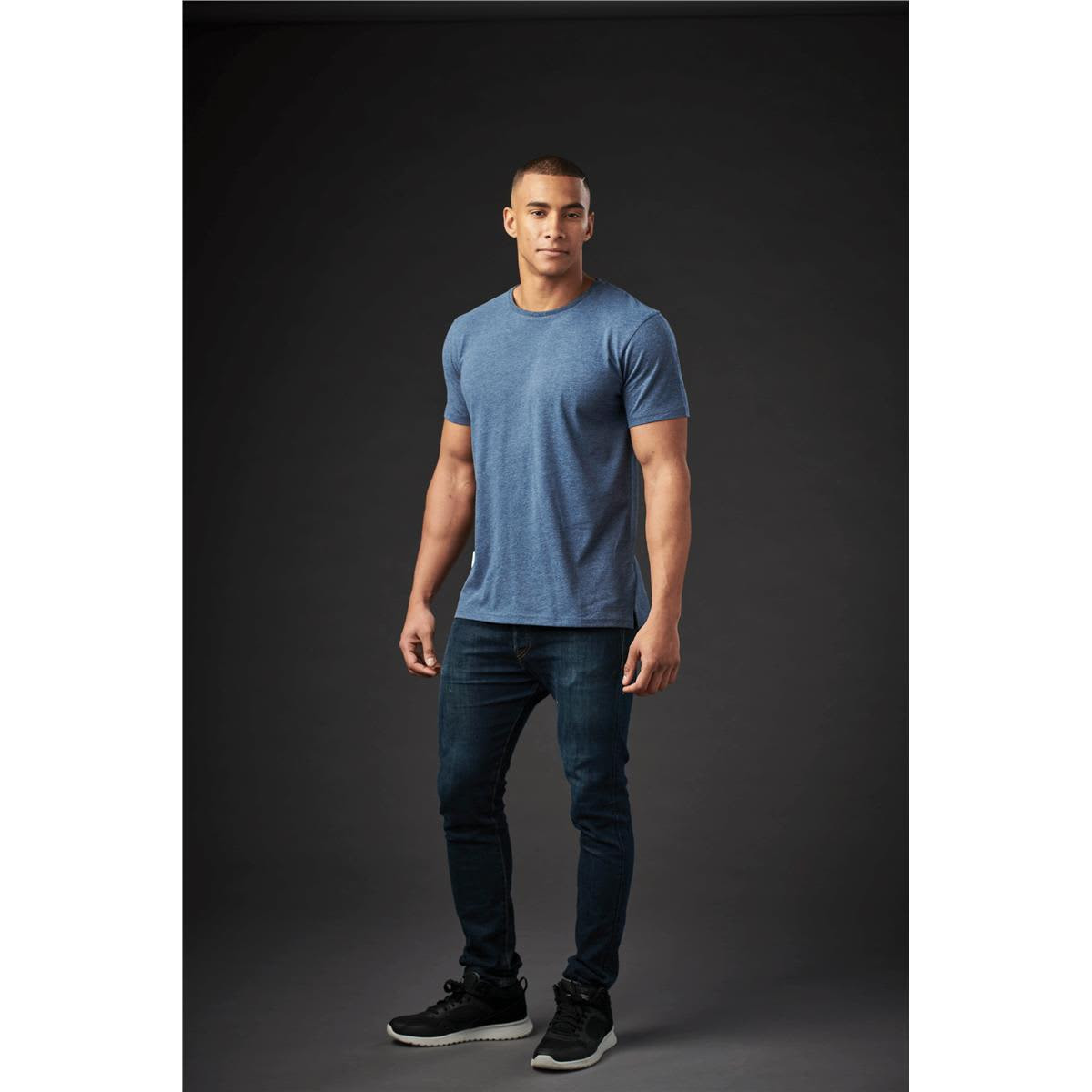A man models a blue crew neck T-shirt and dark jeans, standing against a dark background. The shirt features a logo.