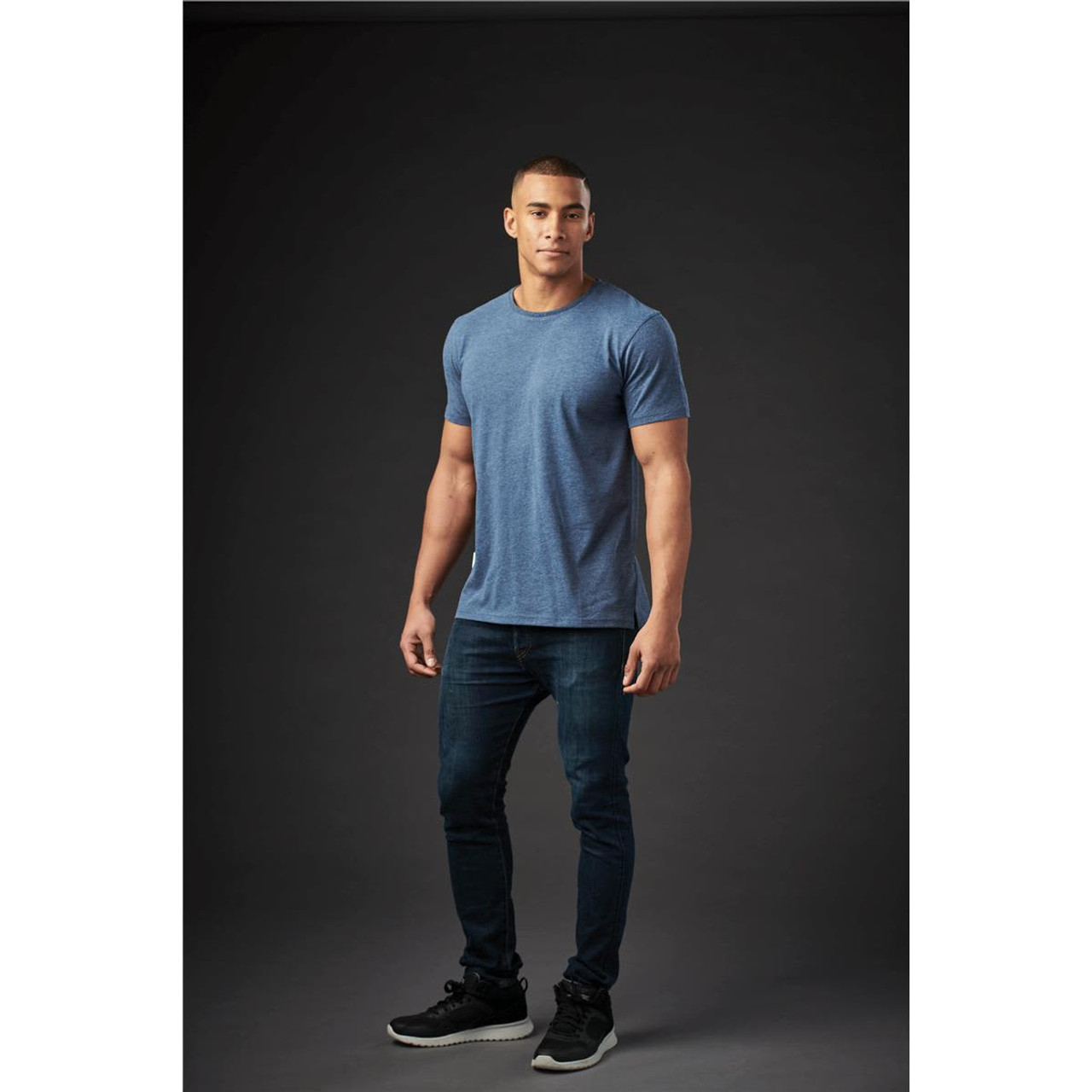 A man models a blue crew neck T-shirt and dark jeans, standing against a dark background. The shirt features a logo.