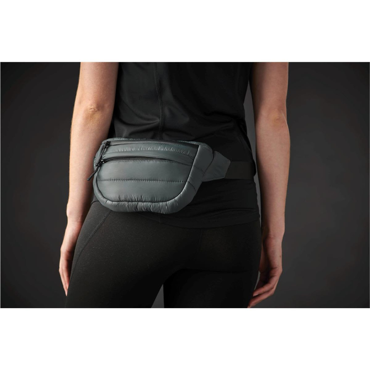 A quilted grey waist bag with a black adjustable strap worn at the waist. The bag features a zipper and a logo.