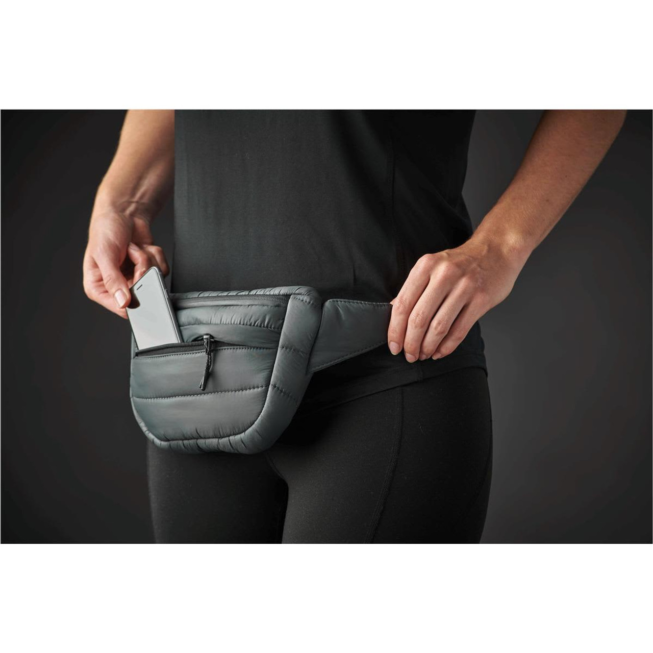 A quilted, dark green waist bag with a small front pocket, worn around the waist, holding a smartphone.