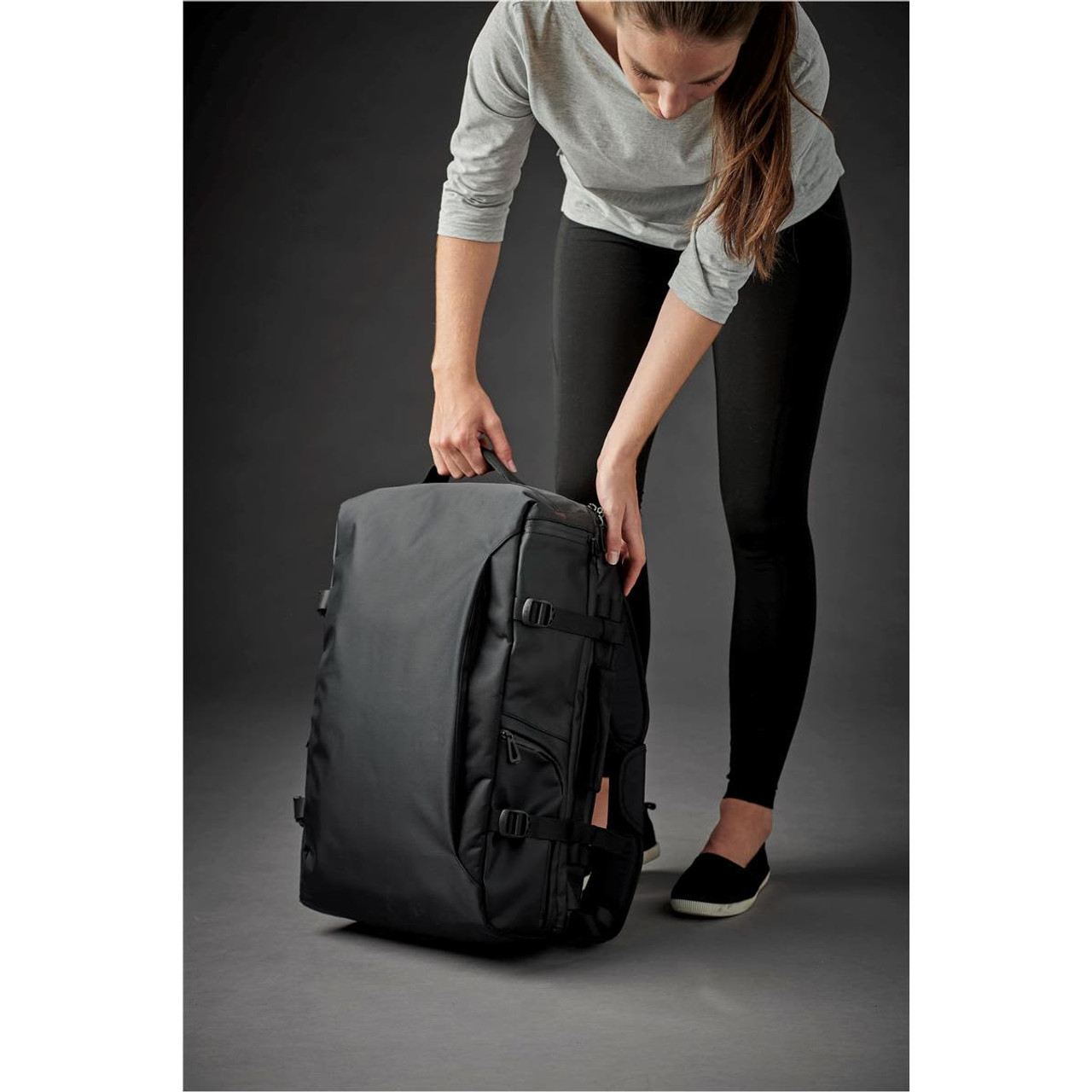 A black backpack being handled by a person, featuring straps and a sleek design. It has a logo on the front.