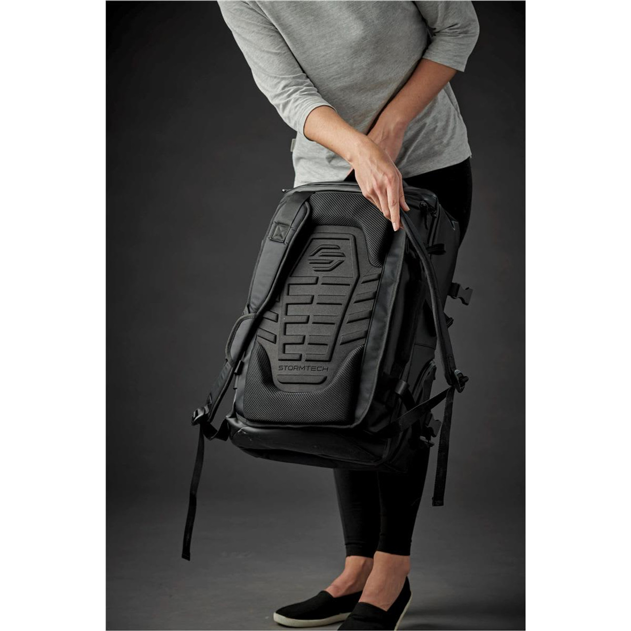 A black backpack with a textured back panel is being held by a person wearing a grey top.