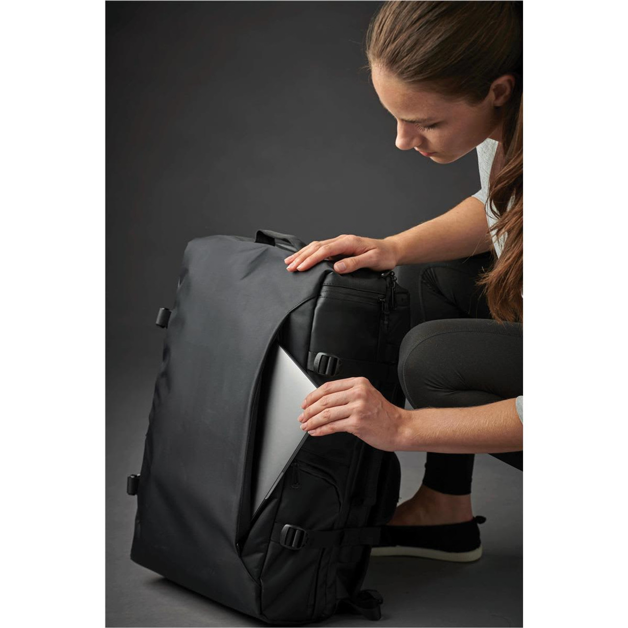 A black backpack with a person inserting a silver laptop into a side pocket. The bag features straps and zippers.