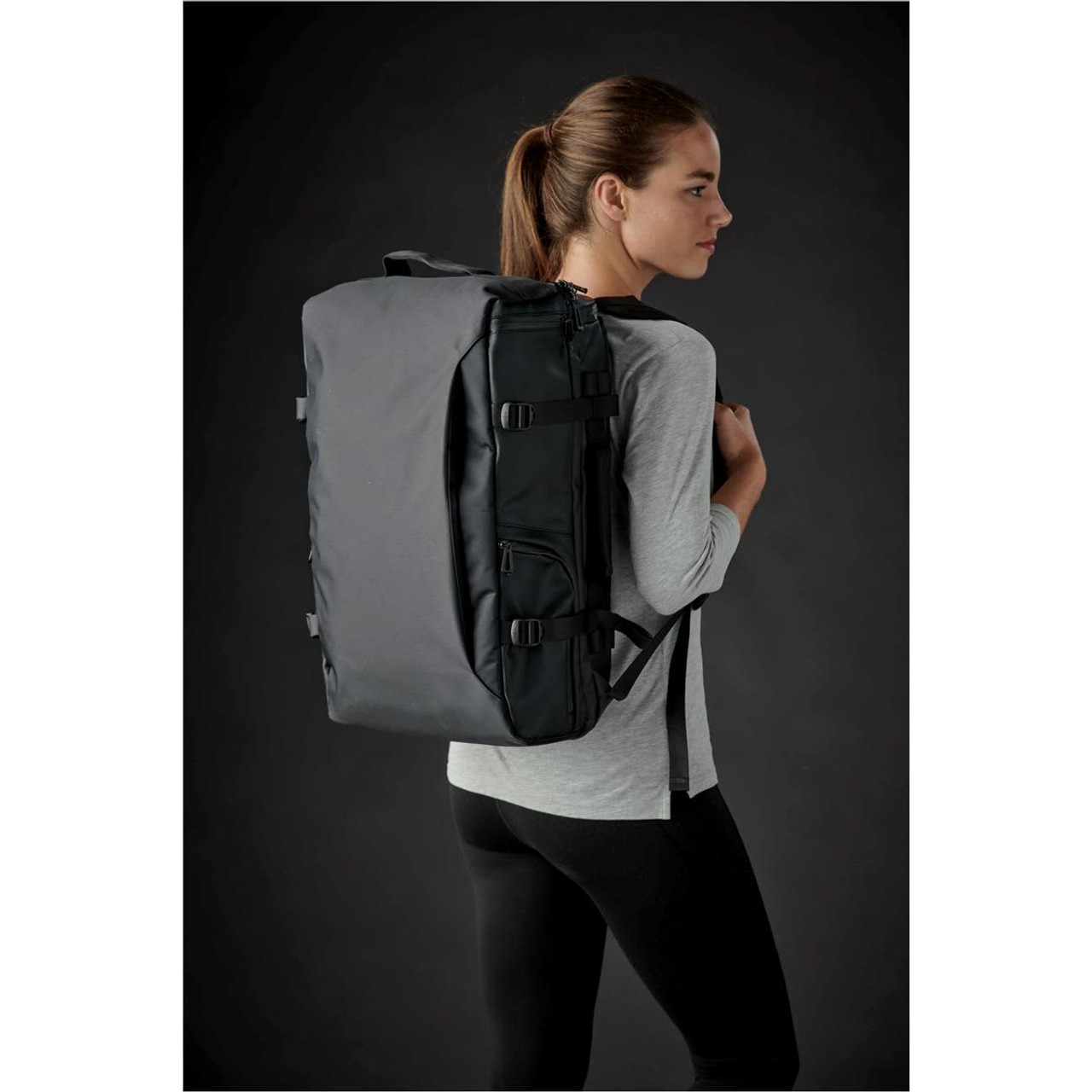 A black backpack with straps, featuring a logo, worn on a model's back against a dark background.