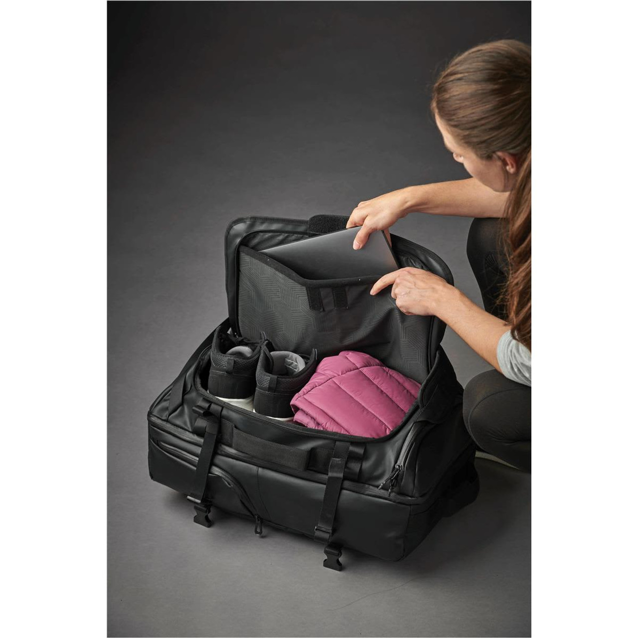 A black backpack with a zippered opening reveals a pink padded item and a pair of shoes, with a laptop sleeve inside.