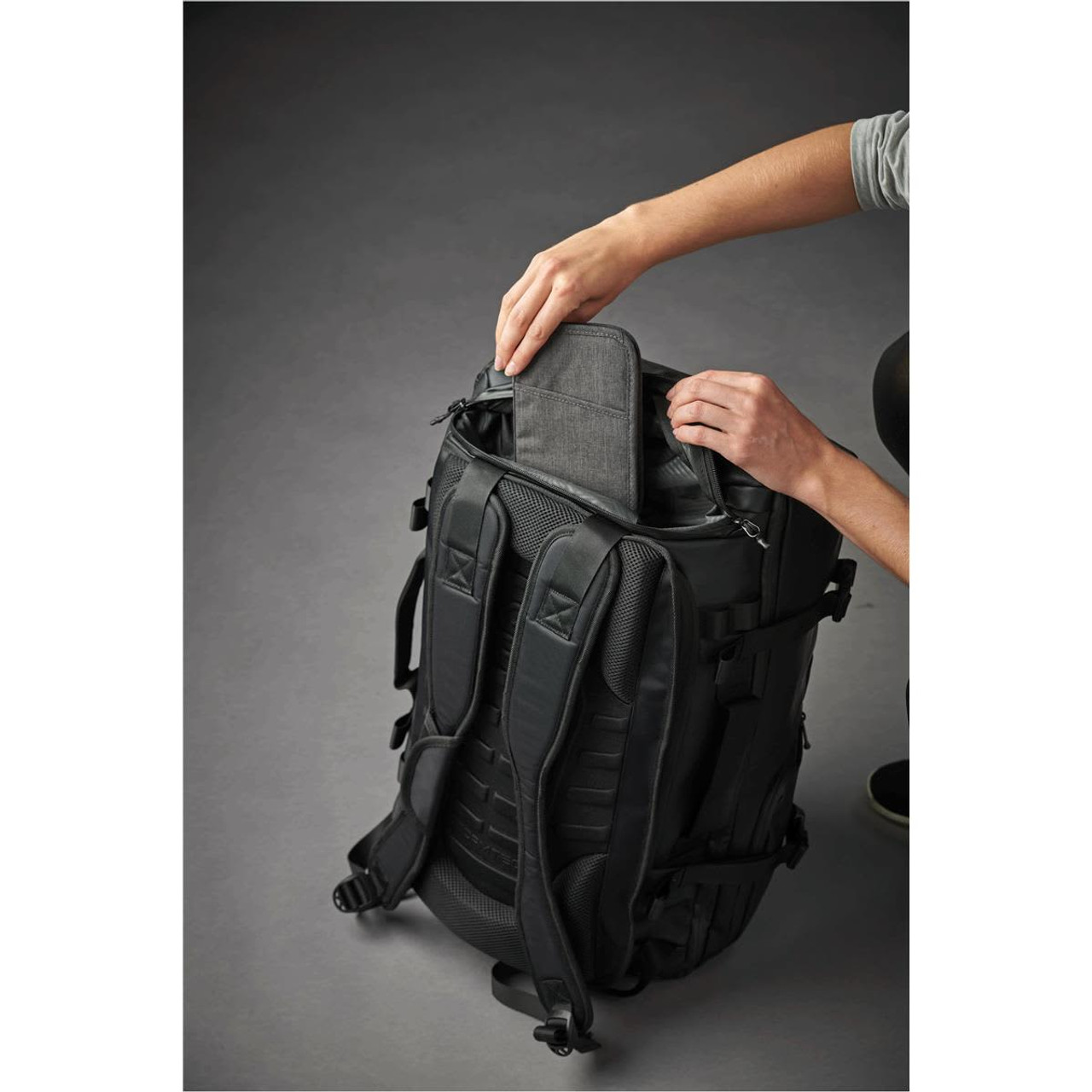 A black backpack with padded straps is being unzipped, revealing a grey compartment at the top. It has a logo.
