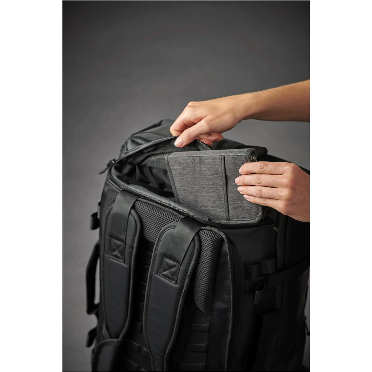 A black backpack with a person placing a grey tablet or book into its top pocket. The backpack features padded straps.