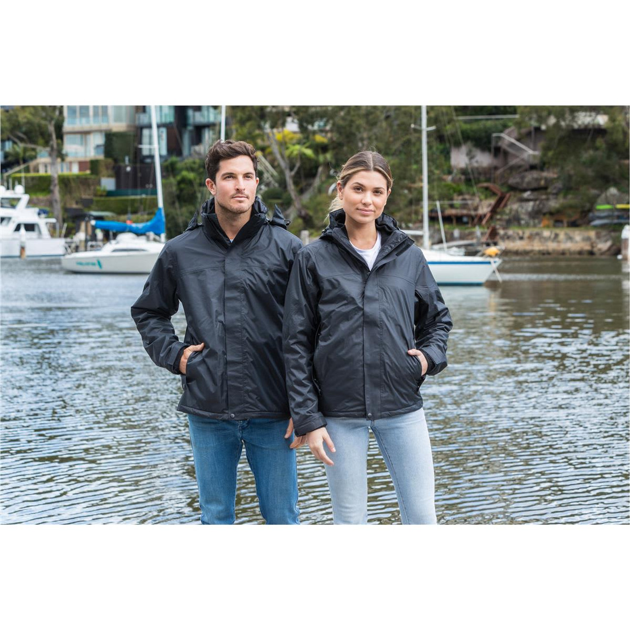 A unisex black 3-in-1 jacket is worn by a man and a woman near a waterfront with boats. The jacket has a logo. A unisex black 3-in-1 jacket is worn by a man and a woman near a waterfront with boats. The jacket has a logo.
