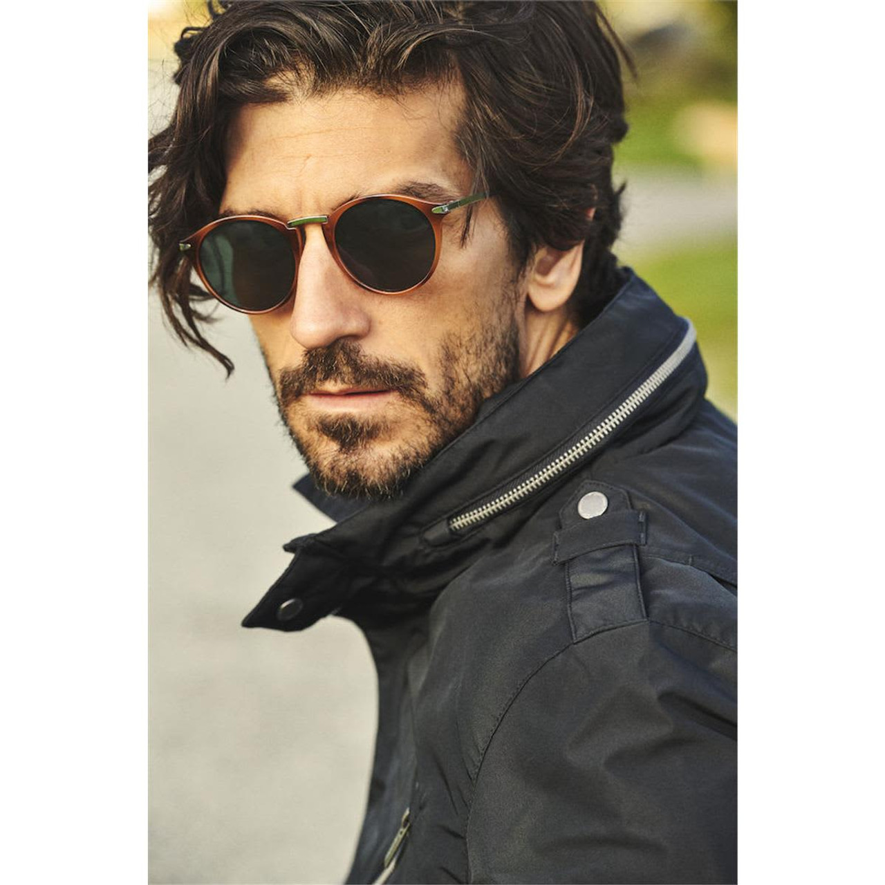 A man wearing a black business coat and round sunglasses, with a stylish hairstyle, outdoors. The coat features a logo.