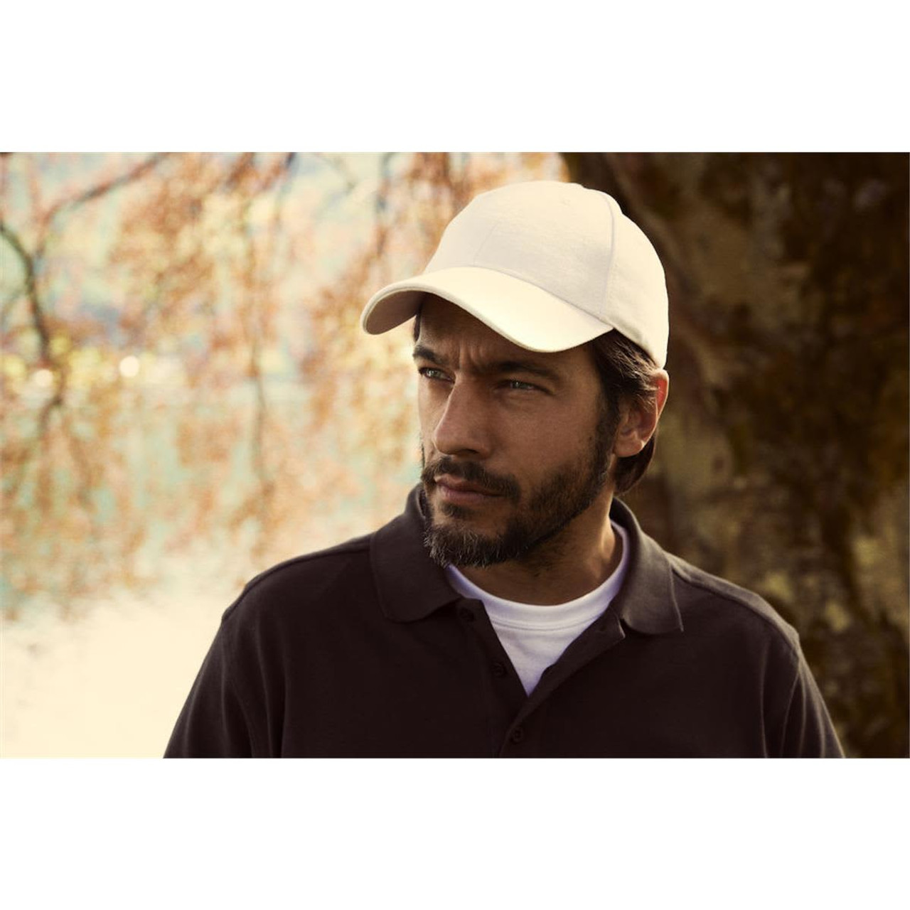 A beige cap worn by a man with a beard, standing outdoors near water and trees. The cap has a logo.