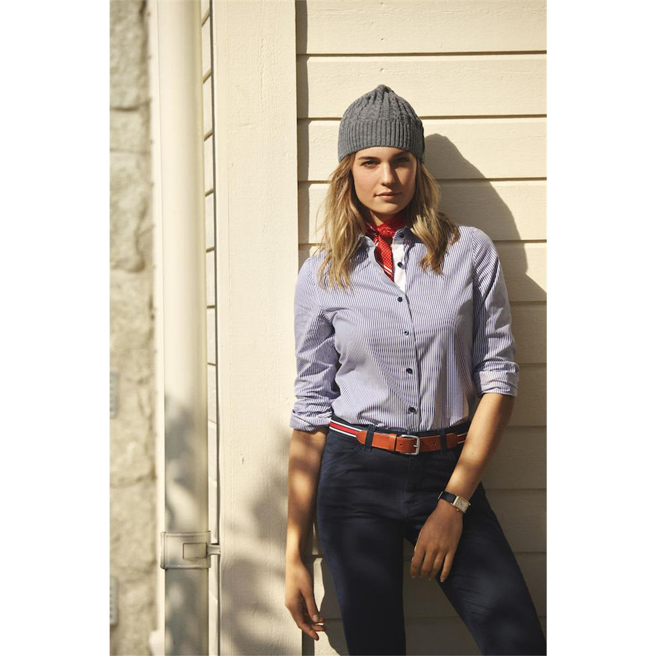 A woman wearing a blue and white striped button-up shirt, styled with a red neck scarf and grey beanie, stands against a wall.