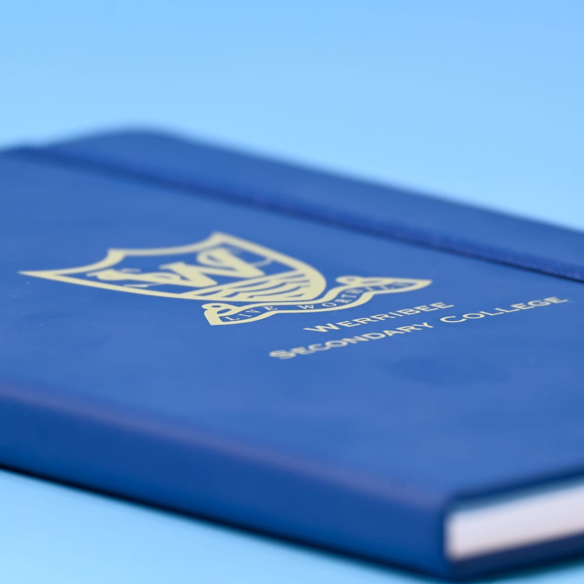 A blue A5 notebook with a logo and gold lettering on the cover, featuring a textured finish.