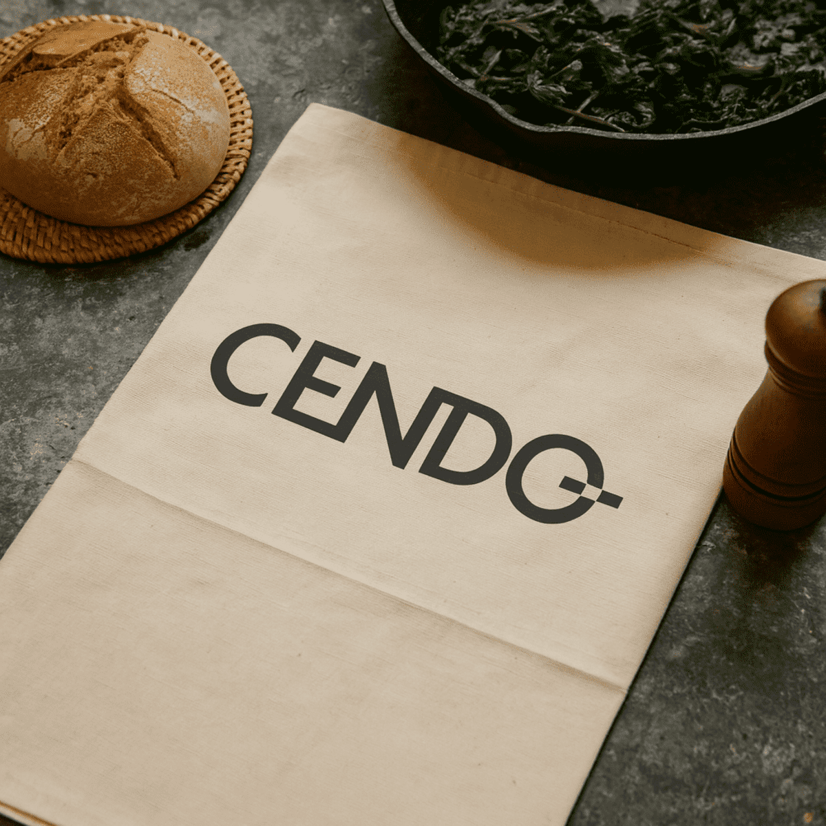 A cotton tea towel in a light cream colour featuring a logo, placed alongside bread and a bowl of greens.