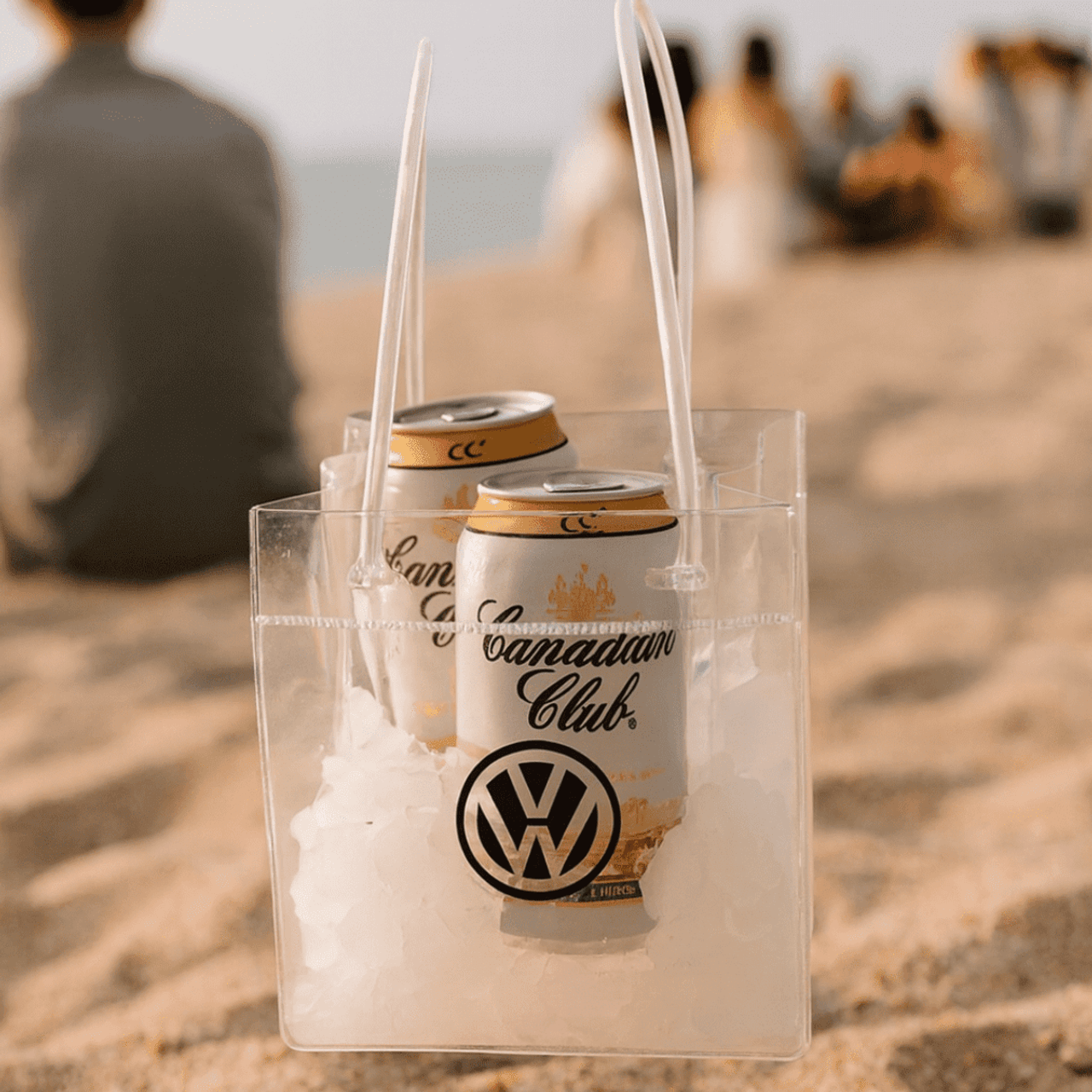A clear cooler bag filled with ice and two cans, featuring a logo. Set on a sandy beach with people in the background.