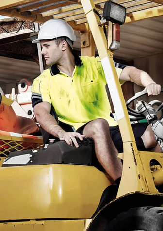 A man wearing a yellow and navy hi-vis short sleeve polo shirt operates a forklift, wearing a safety helmet. A man wearing a yellow and navy hi-vis short sleeve polo shirt operates a forklift, wearing a safety helmet.