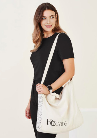 A woman carries a cream unisex tote bag with a logo, wearing a black top and dark trousers.