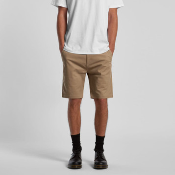 Tan dress shorts displayed on a model wearing a white t-shirt and black shoes. The shorts feature pockets and a logo. Tan dress shorts displayed on a model wearing a white t-shirt and black shoes. The shorts feature pockets and a logo.