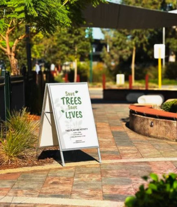 A-frame sign in white and silver displaying a message about saving trees, set in a landscaped outdoor area.