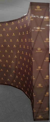 A curved media wall in brown with sides, featuring a repeated logo pattern in gold.