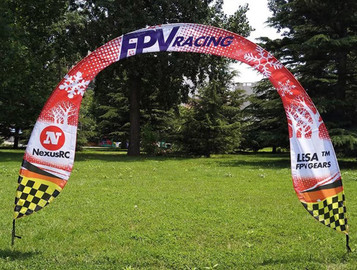An arch banner in red, white, and black with logos, set in a green grassy area surrounded by trees. An arch banner in red, white, and black with logos, set in a green grassy area surrounded by trees.