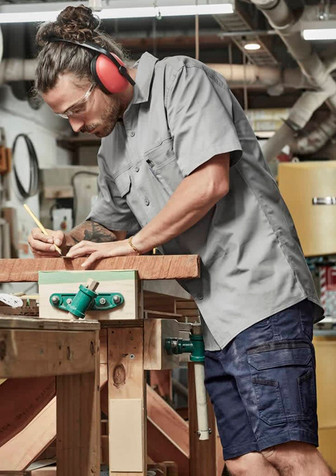A man wears a grey short sleeve shirt with a logo, using a pencil at a workbench in a workshop setting. A man wears a grey short sleeve shirt with a logo, using a pencil at a workbench in a workshop setting.