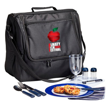 A black picnic basket set includes utensils, a drink glass, condiment containers, and a blue plate with food. A black picnic basket set includes utensils, a drink glass, condiment containers, and a blue plate with food.