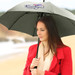 A grey umbrella with a logo, held by a woman wearing a red coat, set against a beach backdrop. A grey umbrella with a logo, held by a woman wearing a red coat, set against a beach backdrop.
