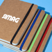 A stack of brown notebooks with colourful elastic bands in blue, green, and red, featuring a logo on the front.