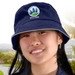 A white bucket hat with a navy sandwich trim and a logo, worn by a smiling person outdoors.