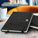 A black notebook with a logo on the cover, resting on a wooden surface near a cup of coffee.