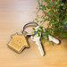 A house-shaped key ring in wood finish, alongside two silver keys and a green plant. It features a brand logo. A house-shaped key ring in wood finish, alongside two silver keys and a green plant. It features a brand logo.