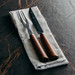 A cheese carving set featuring a knife and fork with wooden handles, positioned on a linen cloth. A cheese carving set featuring a knife and fork with wooden handles, positioned on a linen cloth.