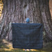 A black puffer blanket is held up by a person in front of a large tree. The scene is outdoors. A black puffer blanket is held up by a person in front of a large tree. The scene is outdoors.