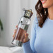 A drink bottle with a stainless steel lid, featuring clear glass and a green logo. The bottle is held by a woman in a blue top. A drink bottle with a stainless steel lid, featuring clear glass and a green logo. The bottle is held by a woman in a blue top.