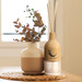 A wooden reed diffuser with light wood finish and three reeds beside a cream and terracotta vase with dried foliage.