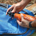A drink bottle in orange with a black spout is being placed onto a blue backpack on a rocky surface.
