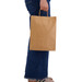 A large brown paper bag held by a person wearing blue jeans and casual shoes. The bag has handles and a simple design. A large brown paper bag held by a person wearing blue jeans and casual shoes. The bag has handles and a simple design.