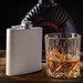 A stainless steel hip flask beside a glass filled with amber liquid and ice, set against a dark background. A stainless steel hip flask beside a glass filled with amber liquid and ice, set against a dark background.