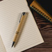 A pen with a brown paper body and silver accents rests on a lined notepad with a spiral notebook nearby. A pen with a brown paper body and silver accents rests on a lined notepad with a spiral notebook nearby.