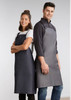 Two models wear Urban Bib Aprons in dark grey and charcoal, featuring pockets and a logo. Two models wear Urban Bib Aprons in dark grey and charcoal, featuring pockets and a logo.