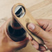 A drink bottle opener made of wood and metal, being used to open a dark beverage bottle. It features a logo. A drink bottle opener made of wood and metal, being used to open a dark beverage bottle. It features a logo.