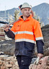 Hi-vis orange and navy waterproof jacket with reflective stripes, worn by a worker holding a tool on a construction site. Hi-vis orange and navy waterproof jacket with reflective stripes, worn by a worker holding a tool on a construction site.