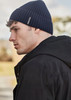 A side view of a man wearing a navy ribbed beanie, paired with a black jacket, outdoors. The beanie features a logo.
